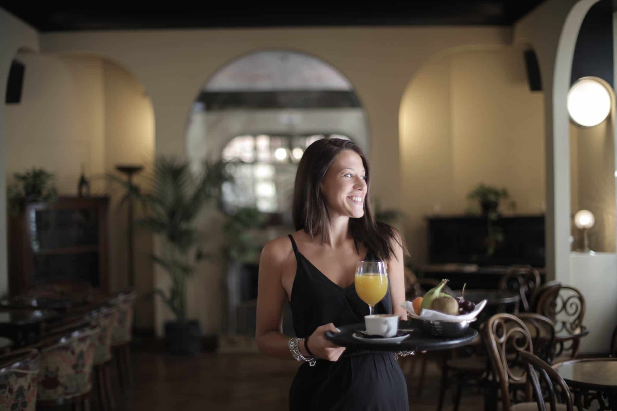 A smiling woman in a chic black dress carries a tray with a glass of orange juice and a cup of coffee in a well-appointed hotel dining area, embodying the attentive service often praised in positive hotel reviews as opposed to negative hotel reviews.