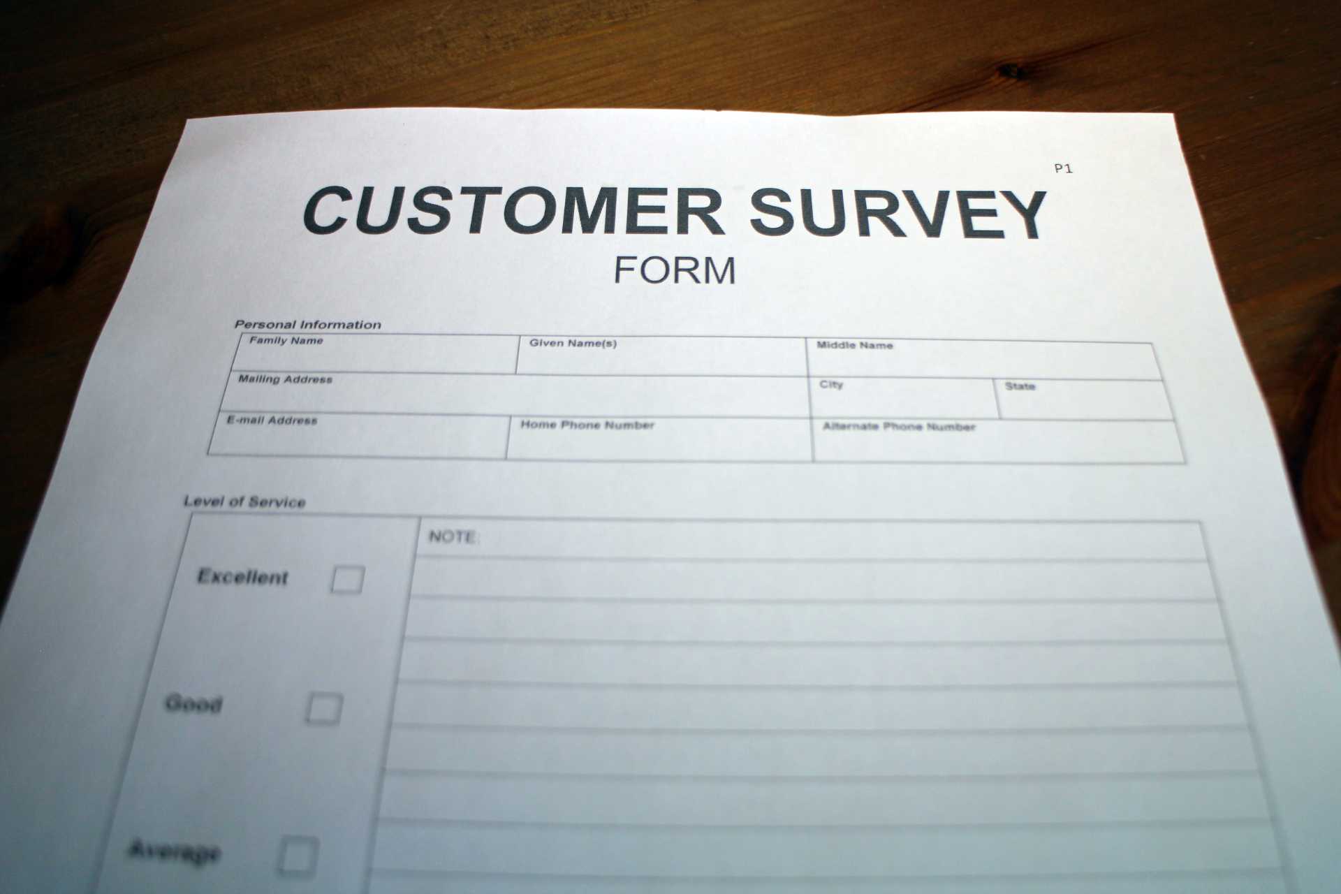 Printed customer survey form on a wooden surface with sections for personal information such as family name, given names, and contact details, followed by a service level rating ranging from 'Excellent' to 'Poor' and a section for additional notes