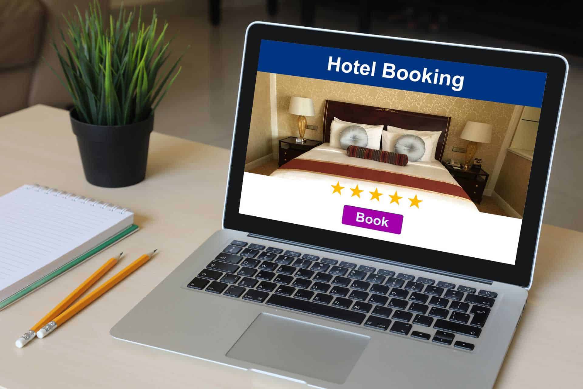 Online hotel booking interface open on a laptop screen with a five-star rating and a 'Book' button, set on a desk with a notebook, pencils, and a potted plant, symbolizing guest intent in the hotel industry towards convenient and rated accommodation options.