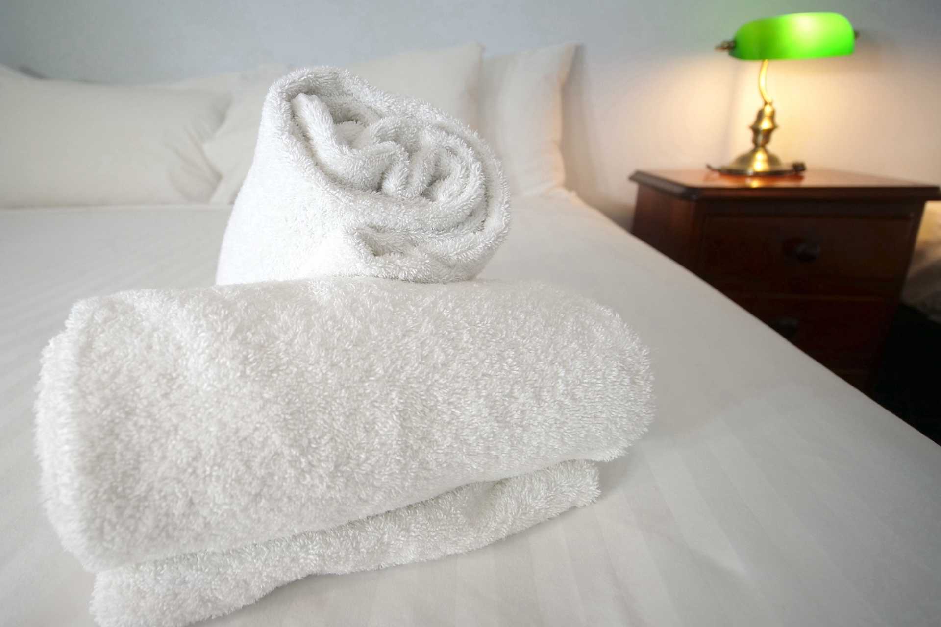 Neatly rolled white towels on a pristine hotel bed with a classic green-shaded bedside lamp in the background, indicative of the meticulous attention to detail that enhances a guest's journey and overall experience in a hotel.